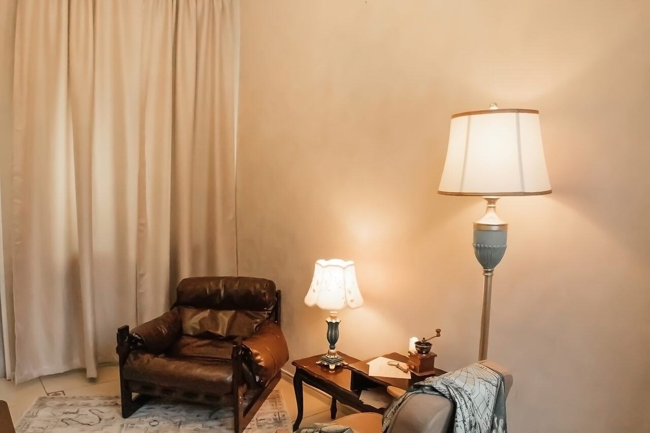 Арендовать Open Space (Открытое пространство) для мероприятий. Гостиная с кожаным креслом, двумя лампами с абажурами на тумбочках, светлыми шторами, светло-бежевыми стенами, ковровым покрытием нейтрального оттенка.