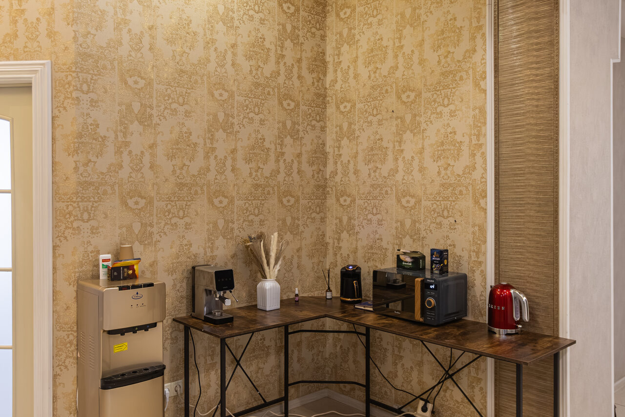 Арендовать Open Space (Открытое пространство) для мероприятий. Угловое пространство с бежевыми узорчатыми обоями. Длинный стол с металлическими ножками. На столе кулер для воды, кофемашина, несколько упаковок кофе, красный электрический чайник. Закрытая дверь слева, освещение мягкое.