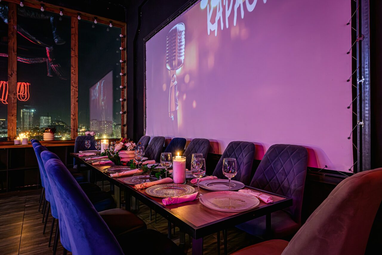 Арендовать Лофт Овертон. Просторное помещение с длинным столом, за которым стоит ряд мягких стульев. На столе сервировка с тарелками и бокалами, украшение из свечей и цветов. На стене большой экран с изображением микрофона. Темное деревянное покрытие пола, окно с видом на ночной город.