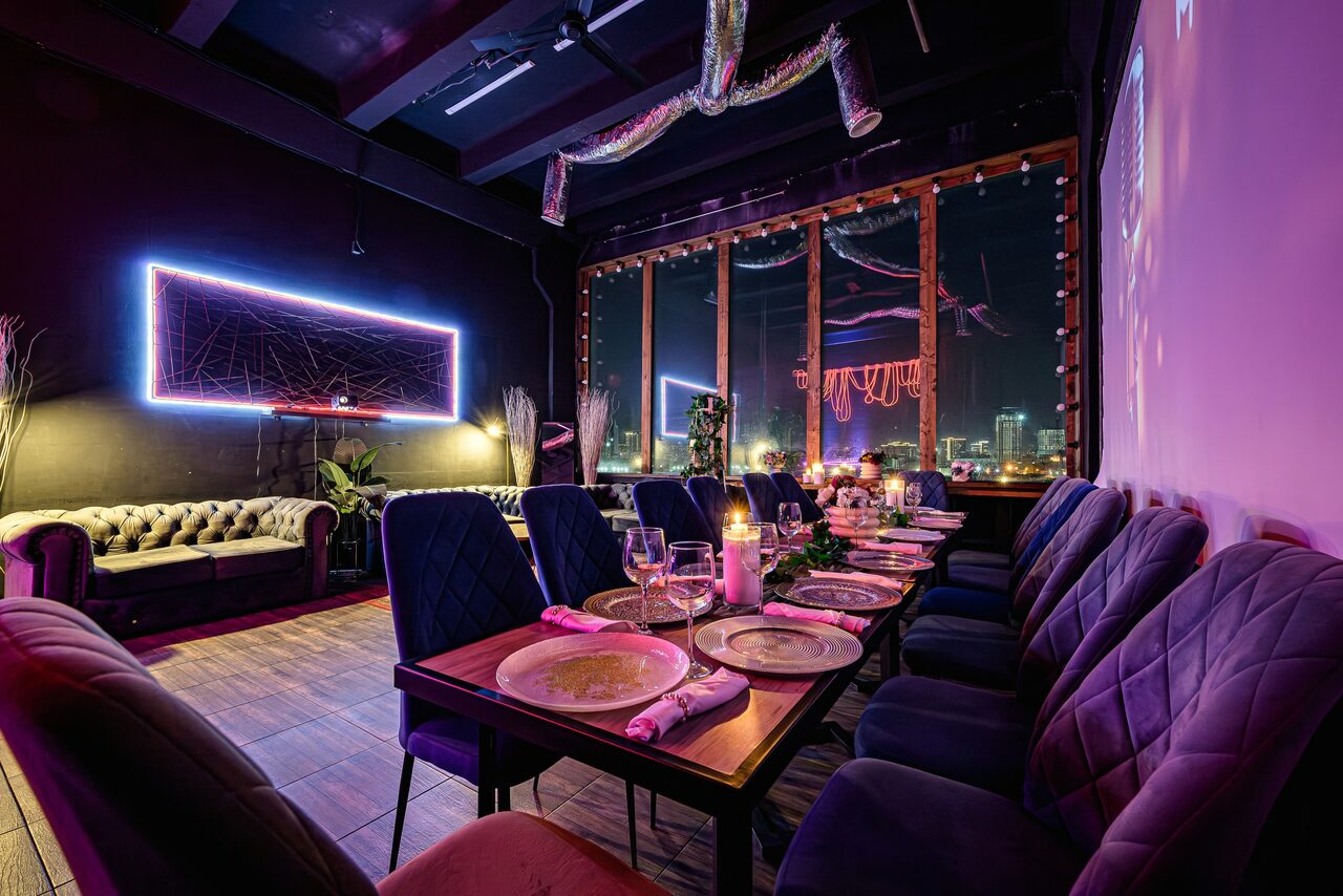 Арендовать Лофт Овертон. Лофт с деревянным полом, длинным столом, сервированным тарелками, бокалами и свечами. Вдоль стола мягкие стулья. На стене неоновая рамка. Большие окна с видом на город. Потолок с декоративными элементами. Освещение разноцветное.