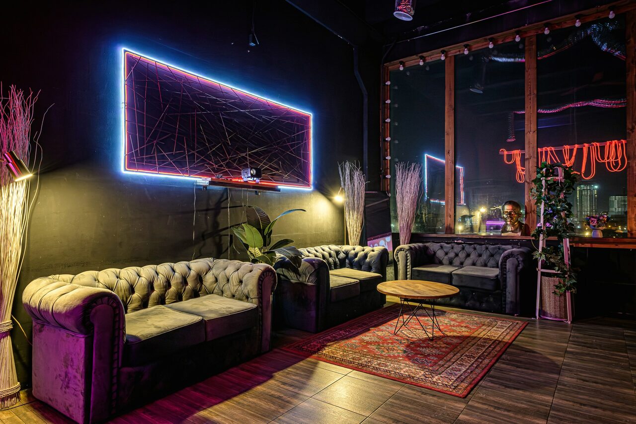 Арендовать Лофт Овертон. Лофт-пространство с темными стенами и неоновым освещением. Расположены три кожаных дивана серого цвета, стеклянный столик, деревянный пол, ковер. Витринное окно с видом на город, растительность, декоративные элементы. Светодиодная панель на стене.