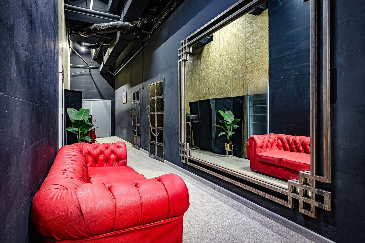 Арендовать Лофт Овертон. Коридор с черными стенами и большим настенным зеркалом, красный диван, искусственные растения в горшках, потолочные трубы, светлая напольная плитка.