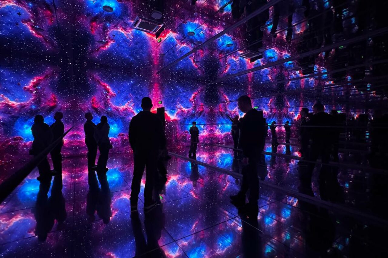 Арендовать Futurione(Футурио́не). Зеркальная комната с космическими проекциями на стенах и потолке, c яркими фиолетовыми и голубыми цветами. На полу зеркальная поверхность, отражающая изображение. Несколько людей стоят и смотрят вокруг. Освещение плавное и многоцветное.