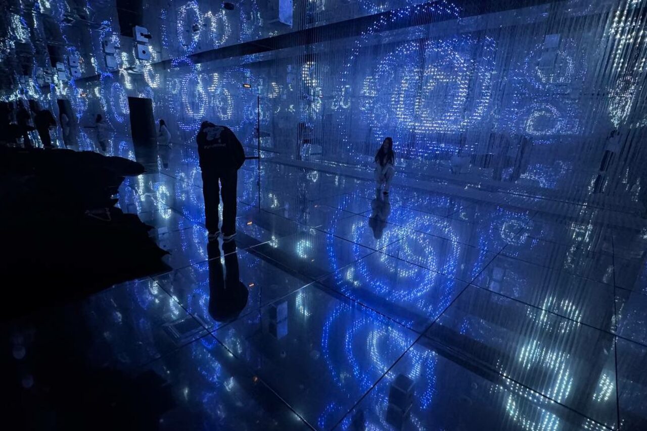 Арендовать Futurione(Футурио́не). Темное помещение с отражающим полом, стенами с нависающими нитями, подсвеченными синим светом. На полу и стенах видны узоры в виде спиралей. Несколько человек в пространстве, отражаются на полу.