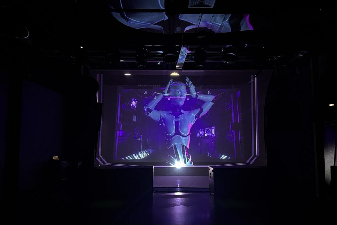 Арендовать Futurione(Футурио́не). Тёмное пространство с большой видеопроекцией, изображающей робота на экране с фиолетовым освещением. Чёрные стены и потолок. На потолке видны световые приборы и конструкции. Пол также тёмный.