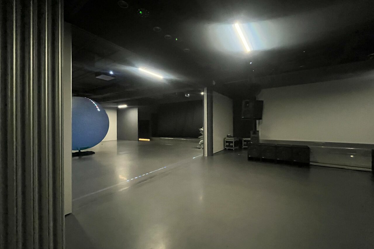 Арендовать Futurione(Футурио́не). Просторное помещение с гладким серым полом, темными стенами и потолком, освещенное потолочными светильниками. Слева виден большой синий объект. Справа расположено звуковое оборудование и черные диваны.