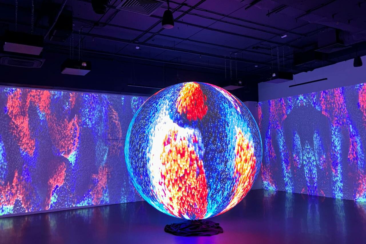 Арендовать Futurione(Футурио́не). Тёмное помещение с бетонным полом и высокими потолками. В центре - сферическая инсталляция с яркой абстрактной проекцией. Стены также покрыты абстрактными проекциями в синих, оранжевых и красных тонах. Освещение приглушённое.