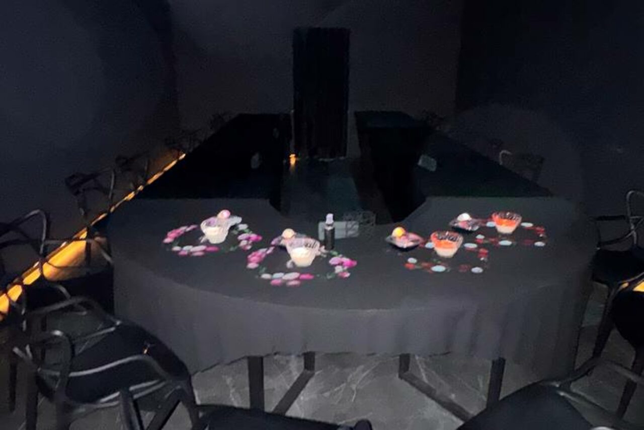 Арендовать Futurione(Футурио́не). Арендуемое пространство с тёмными стенами и обстановкой. Центральный стол в форме буквы "П" с тёмной скатертью, украшенный лепестками цветов и чашами с закусками. Вокруг стола расставлены стулья. Освещение мягкое, неяркое.