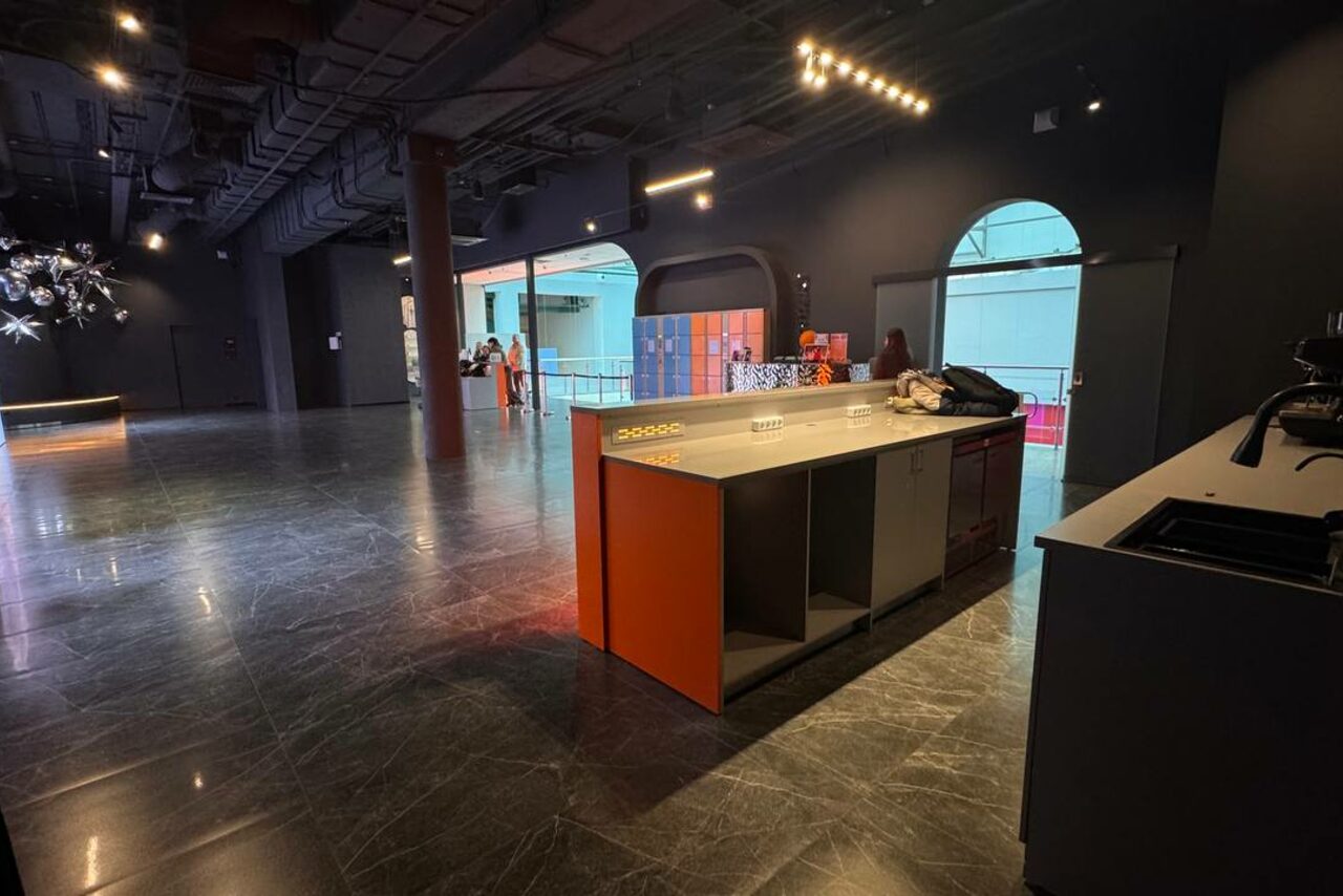 Арендовать Futurione(Футурио́не). Просторное помещение с бетонным потолком, черными стенами и металлическими колоннами. На полу темная плитка. Справа стойка с раковиной. В центре большой стол с цветными боковинами. Потолочное освещение. Вдали видны люди и декоративные предметы.