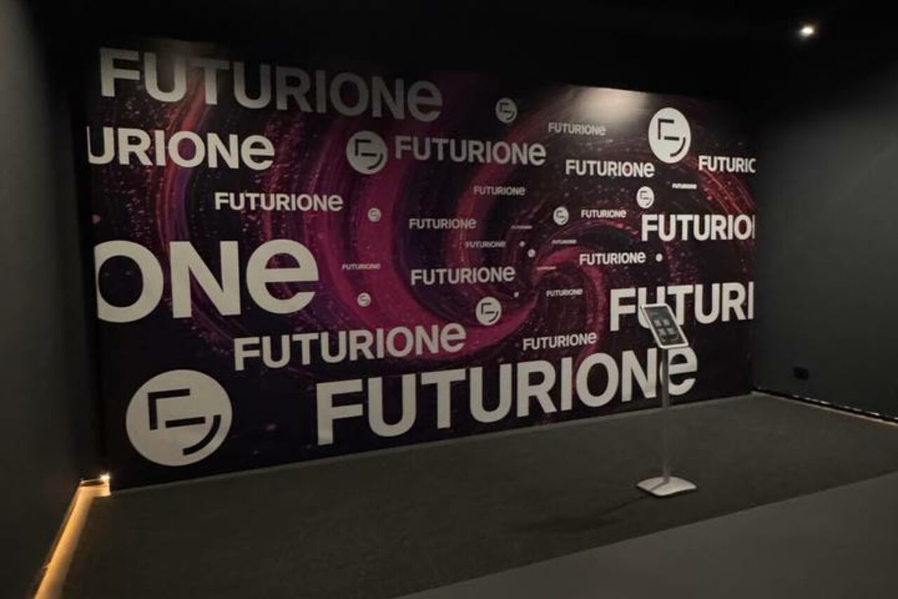 Арендовать Futurione(Футурио́не). Пространство с темными стенами и полом, большая стена украшена фиолетово-белым графическим дизайном с текстом "Futurione". В центре стены планшет на подставке. Освещение мягкое и направленное.