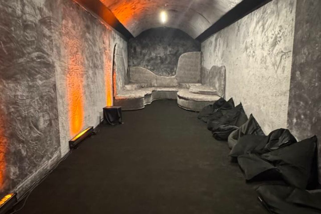 Арендовать Futurione(Футурио́не). Подвал с арочной потолочной конструкцией, серые стены с фактурным узором, черный ковер. Серый угловой диван, черные бескаркасные кресла. Освещение точечными лампами и оранжевой подсветкой по бокам.