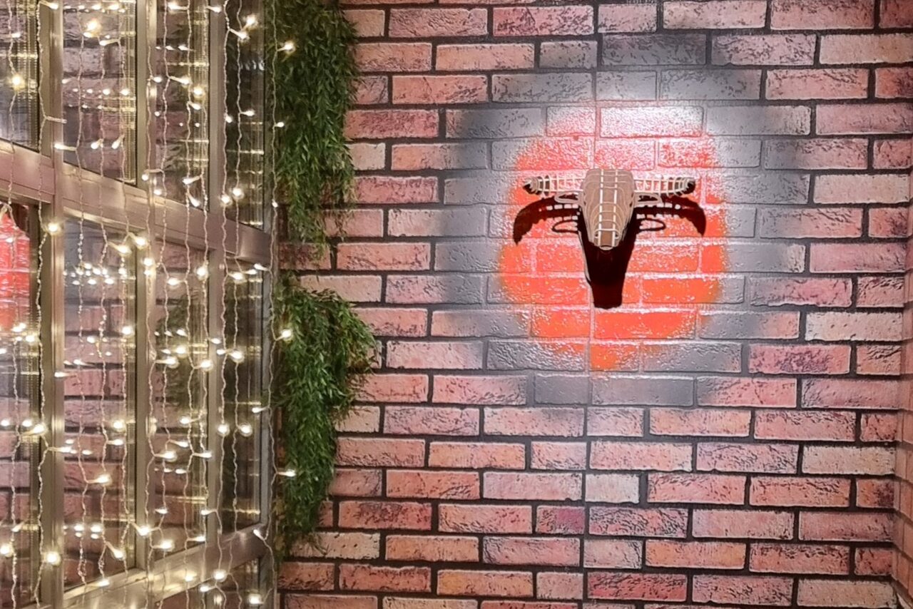 Арендовать BULL LOFT(Лофт Бык) . Кирпичная стена с граффити красного круга и черного рисунка. Слева вертикальная конструкция с растениями и гирляндой светящихся лампочек.