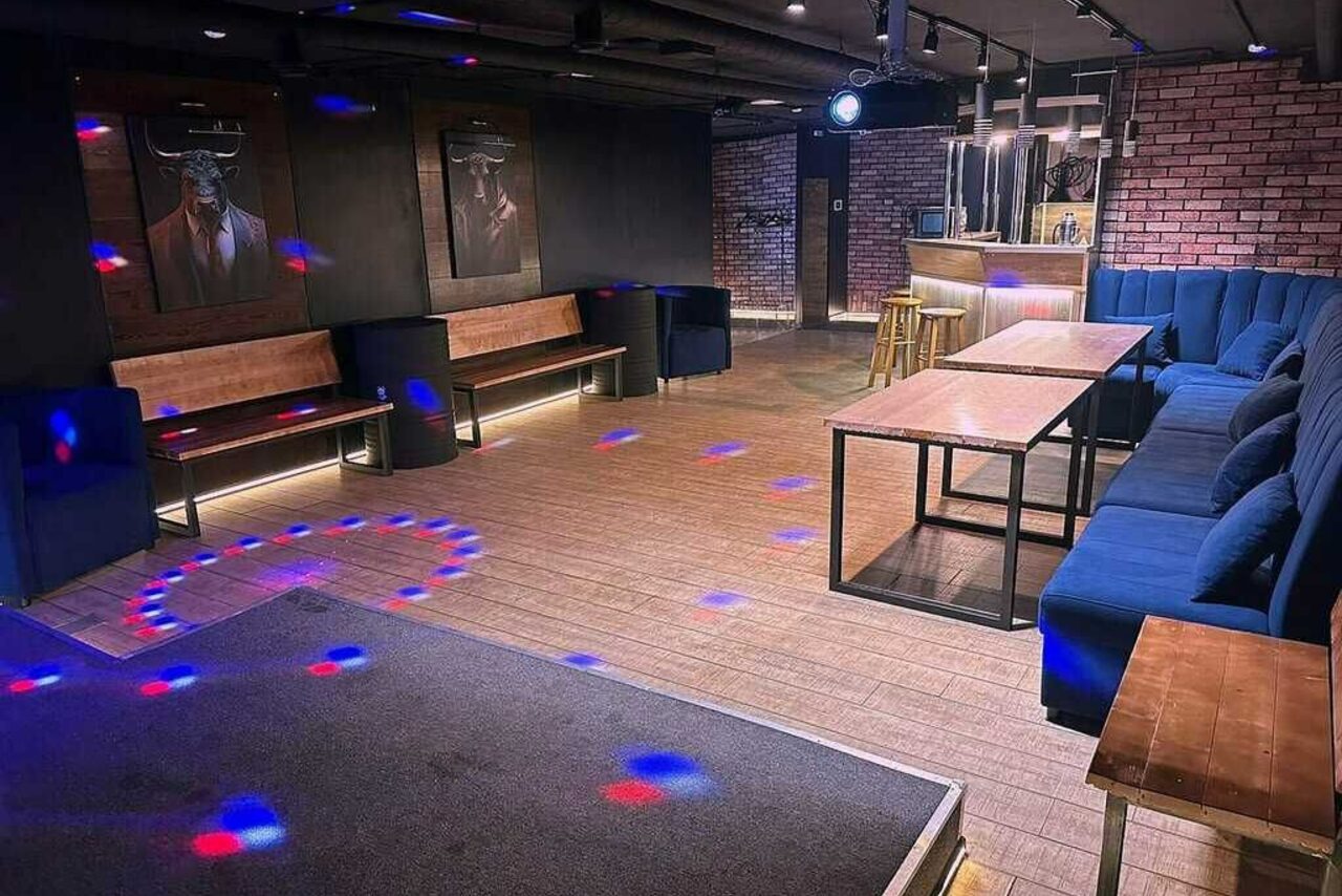 Арендовать BULL LOFT(Лофт Бык) . Лофт-пространство с кирпичными стенами, деревянным полом и баром. Угловой диван и скамьи вдоль стен, длинные деревянные столы. Потолочное освещение, неоновая подсветка и световые пятна на полу. Два постера на стене.