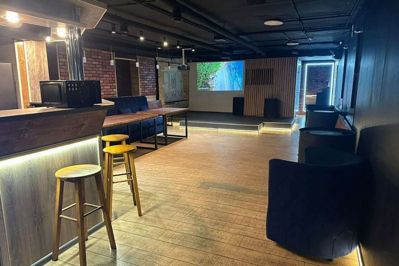 Арендовать BULL LOFT(Лофт Бык) . Современное помещение с деревянными полами и кирпичными стенами. Барная стойка с высокими стульями, черные кресла, проектор с изображением на экране, направленное освещение на потолке, деревянные панели, светлое и просторное пространство.