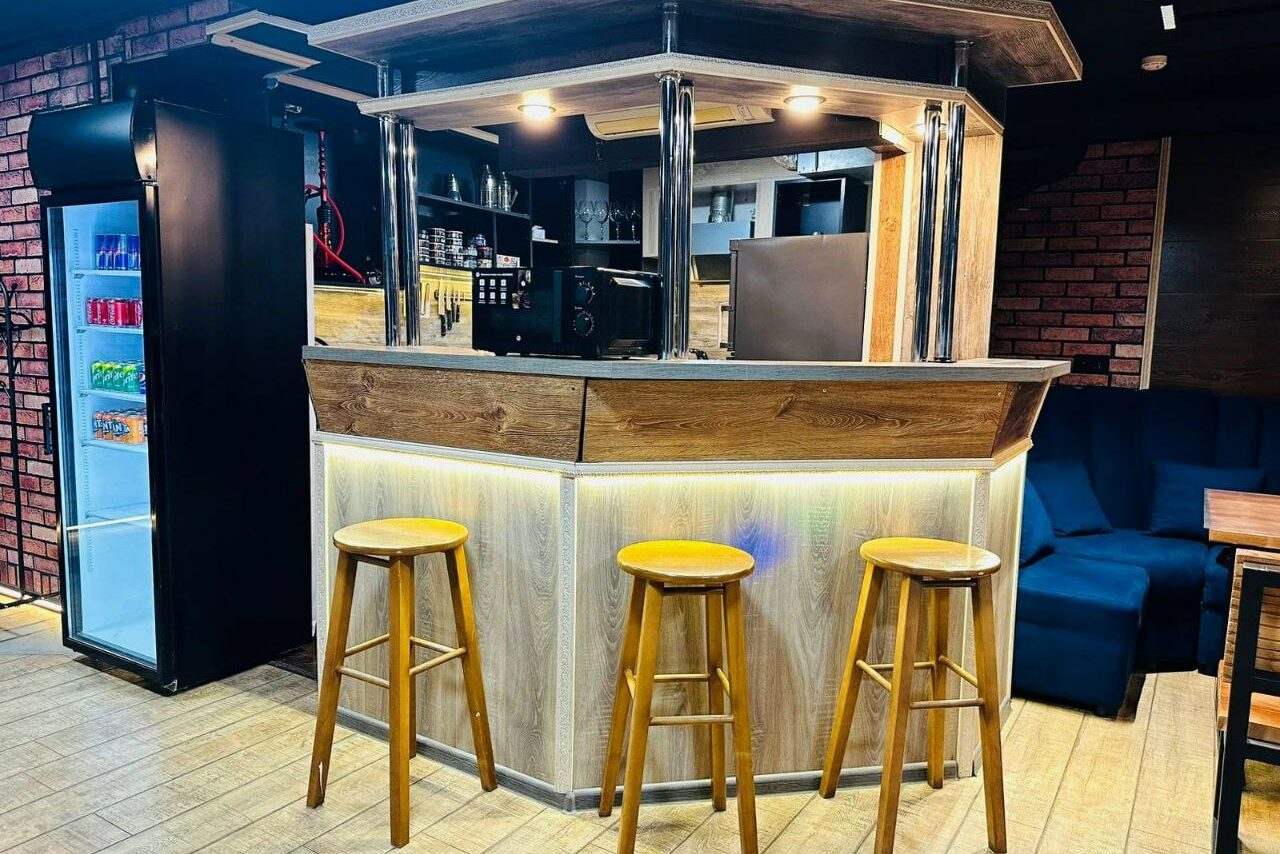 Арендовать BULL LOFT(Лофт Бык) . Барная стойка с деревянной отделкой, три деревянных стула. За стойкой полки с напитками и оборудованием. Справа тёмно-синий диван и деревянный стол. Светлое деревянное покрытие пола, кирпичная стена, холодильник с напитками слева.