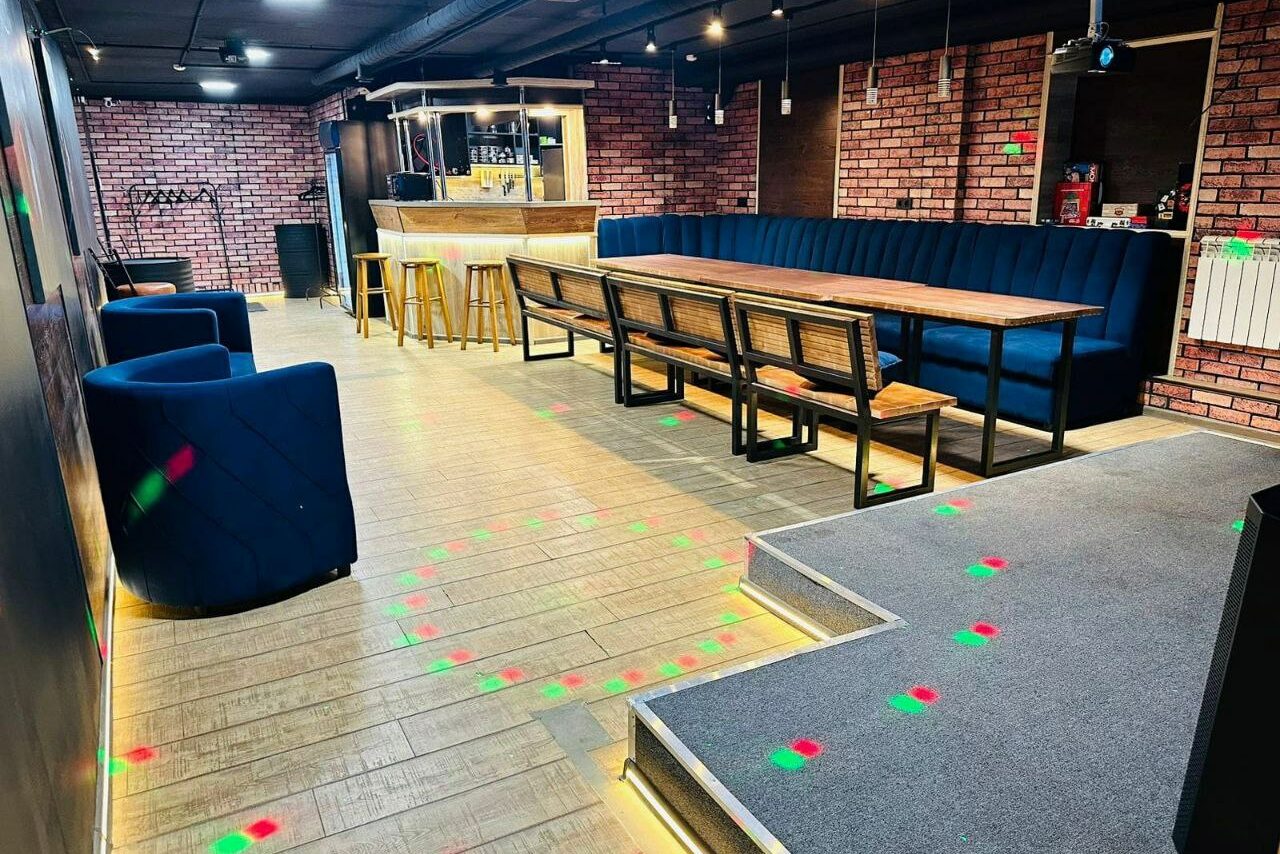 Арендовать BULL LOFT(Лофт Бык) . Арендуемое пространство с барной стойкой; кирпичные стены; деревянные полы и барные стулья; длинный стол с лавкой и синими креслами; черный потолок со светильниками; разноцветные световые точки на полу.