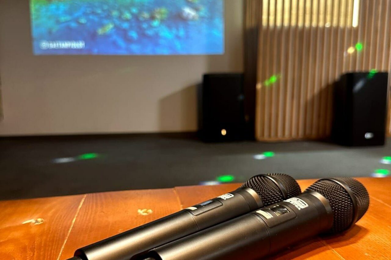 Арендовать BULL LOFT(Лофт Бык) . Пространство с деревянным столом, на котором лежат два микрофона. На заднем плане проектор с изображением на экране. Справа вертикальные жалюзи, на полу световые проекции. Общий цветовой фон теплый.