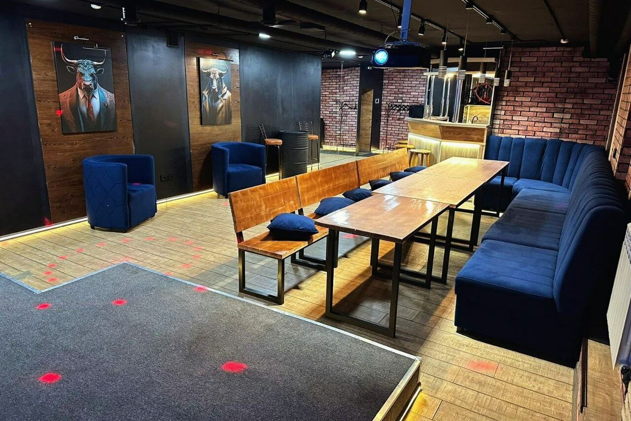 Арендовать BULL LOFT(Лофт Бык) . Просторное помещение с деревянным полом, кирпичными и темными стенами. В центре длинный деревянный стол с лавками и стульями синего цвета. Мягкие кресла, барная стойка. Потолочные светильники, картины на стенах.