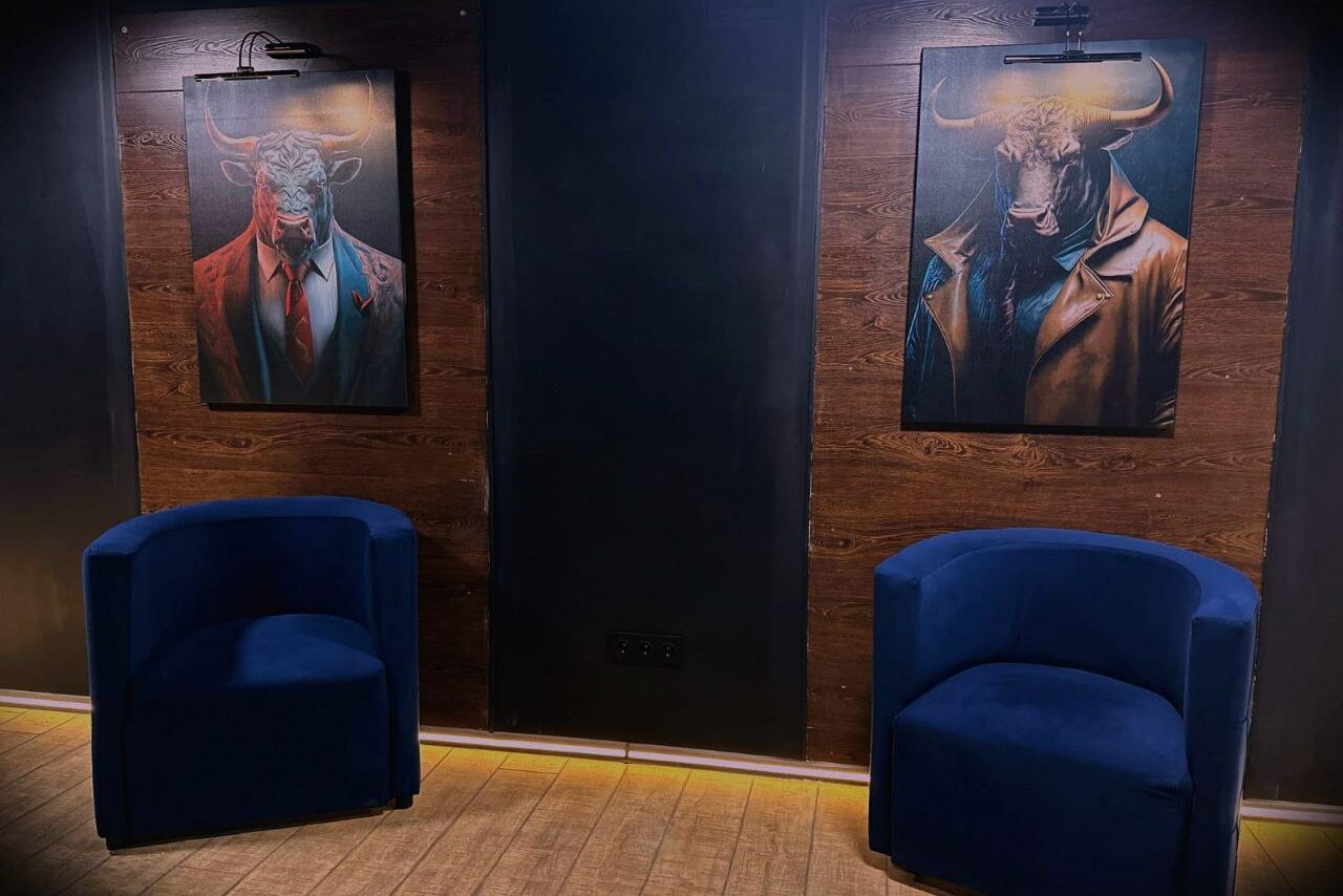 Арендовать BULL LOFT(Лофт Бык) . Два синих кресла на деревянном полу перед стеной с деревянными панелями и черными вставками. На стене висят две картины с изображением быков в одежде, каждая подсвечена лампой.