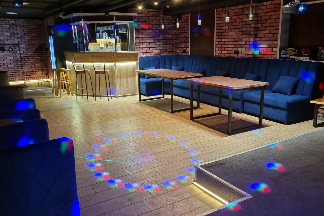 Арендовать BULL LOFT(Лофт Бык) . Пространство с барной стойкой и высокими стульями, рядом столы с синими диванами. Кирпичная стена, деревянный пол. Потолок с точечным освещением. На полу световые проекции.