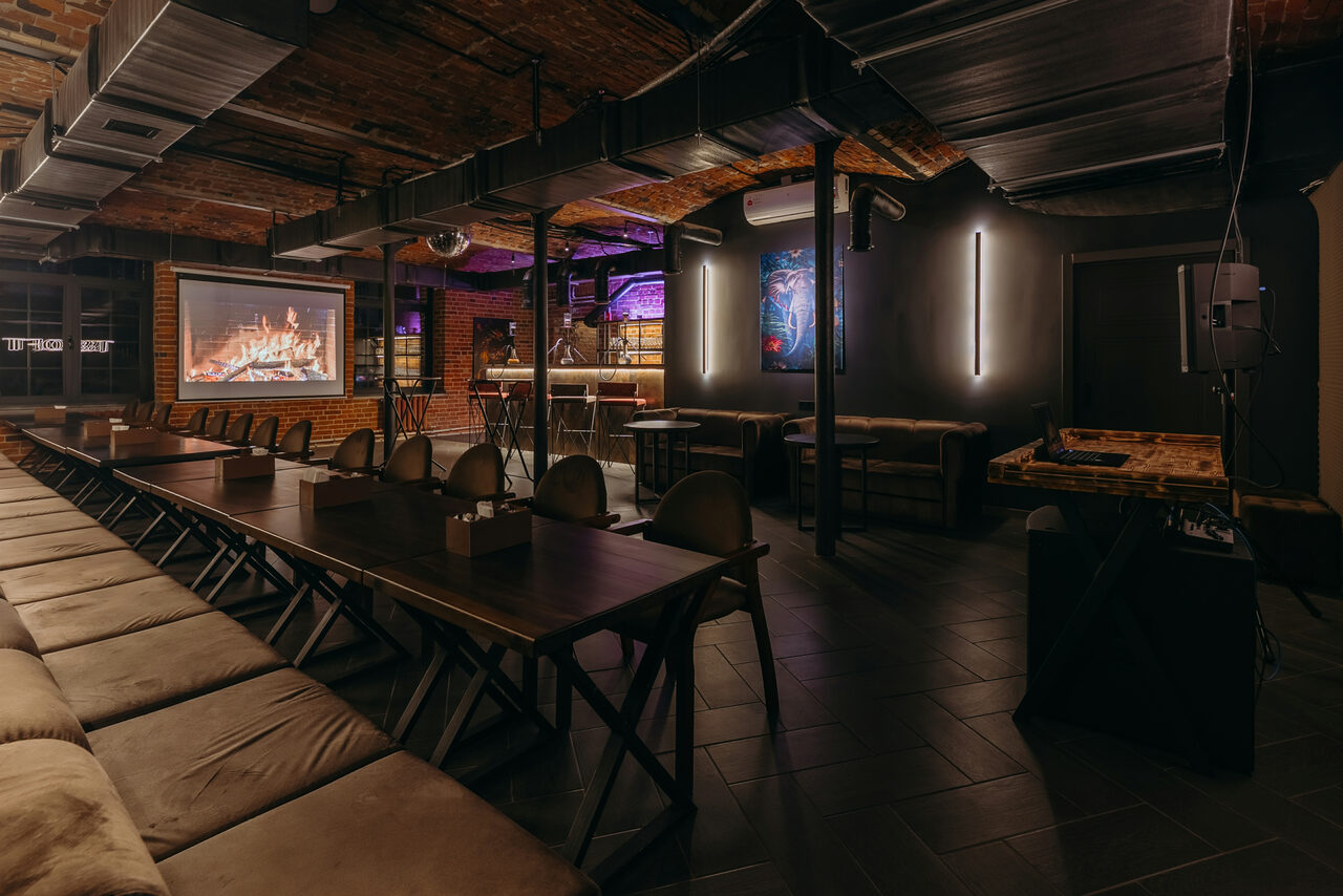 Арендовать Авторские пространство T&S loft (ти энд си лофт) . Лофт-пространство с длинным столом и стульями, кирпичные стены, металлические балки, подвесной светильник, настенная проекция с костром, игровые автоматы, барная стойка, темный кафельный пол, виден неоновый свет, вентиляционные трубы на потолке.