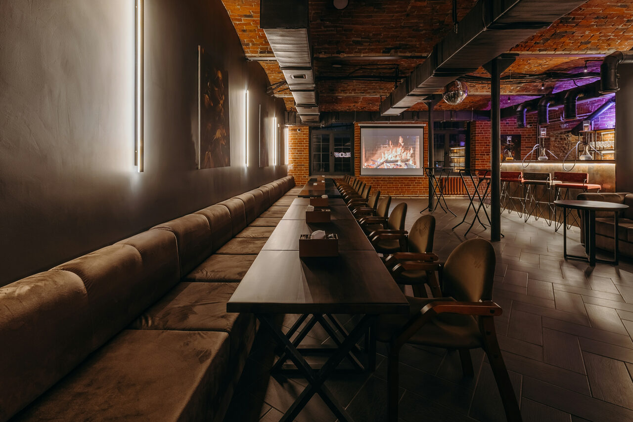 Арендовать Авторские пространство T&S loft (ти энд си лофт) . Темное пространство с кирпичными стенами и потолком. Длинный вдоль стены диван и деревянные столы, окруженные стульями. На противоположной стене проекция камина. Современные бра на стенах, неоновые акценты. Пол выложен плиткой.