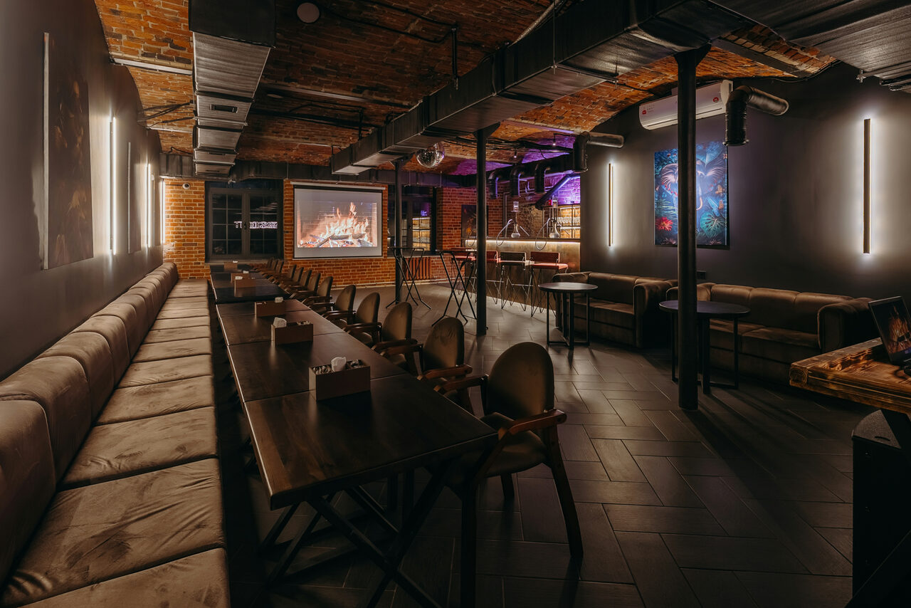 Арендовать Авторские пространство T&S loft (ти энд си лофт) . Лофт-пространство с кирпичными стенами и черным потолком. Есть длинный стол с мягкими креслами и диван вдоль стены. На стене проектор с изображением камина. Освещение светодиодное и неоновое. Пол из темной плитки.