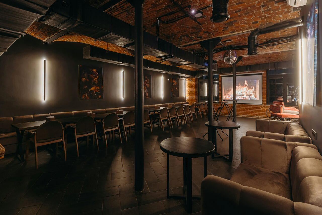 Арендовать Авторские пространство T&S loft (ти энд си лофт) . Подвальное помещение с кирпичными стенами и сводчатым потолком. Зона с диванами, столы и стулья вдоль стены. Вертикальные светильники, проектор с изображением камина. Тёмные тона, металлические колонны, декоративные элементы на стенах.