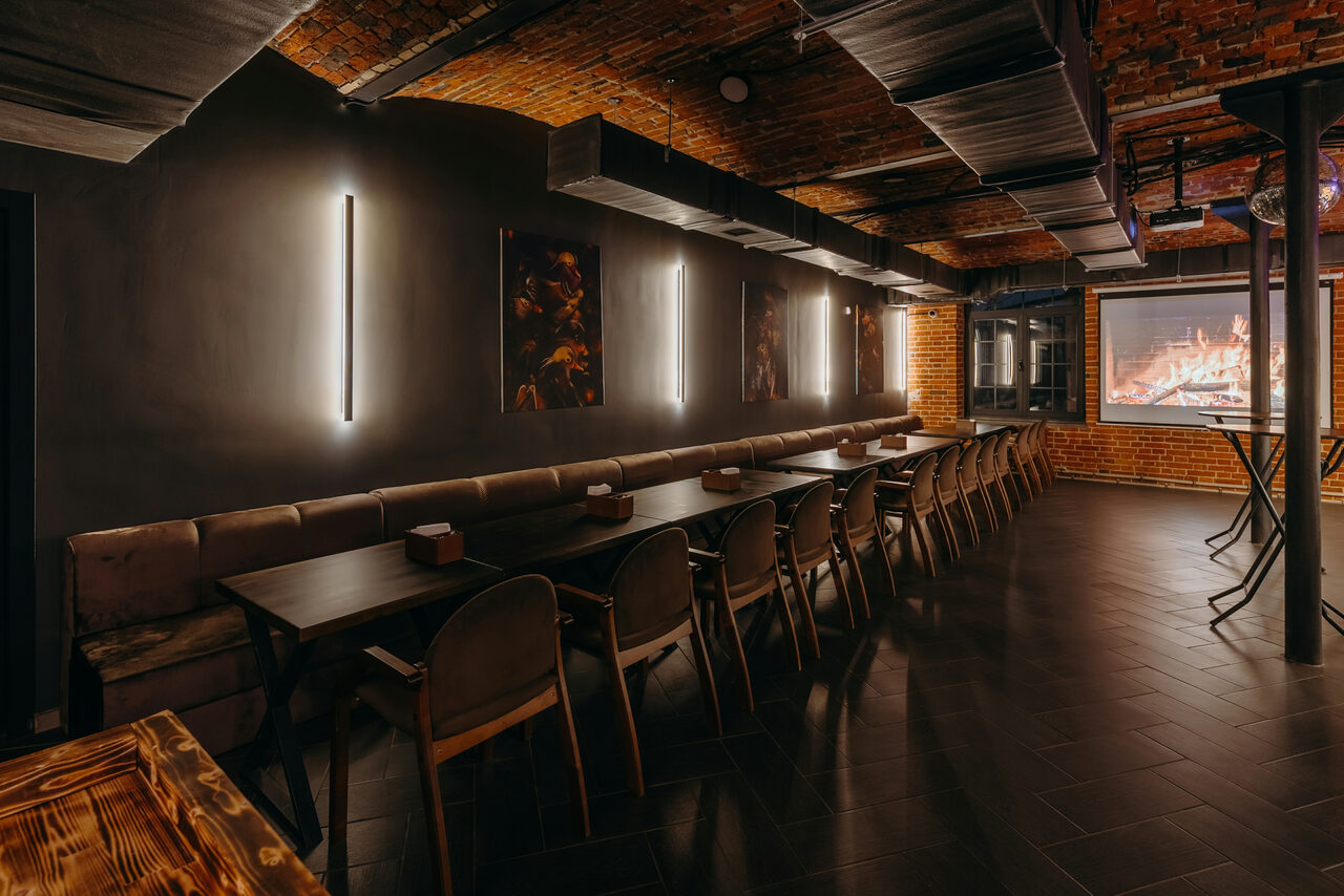 Арендовать Авторские пространство T&S loft (ти энд си лофт) . Просторное помещение с длинным деревянным столом и рядом стульев. Черный матовый потолок с металлическими элементами и кирпичные стены. Светодиодное освещение, картины на стене, большой экран с изображением огня. Темные плиточные полы.
