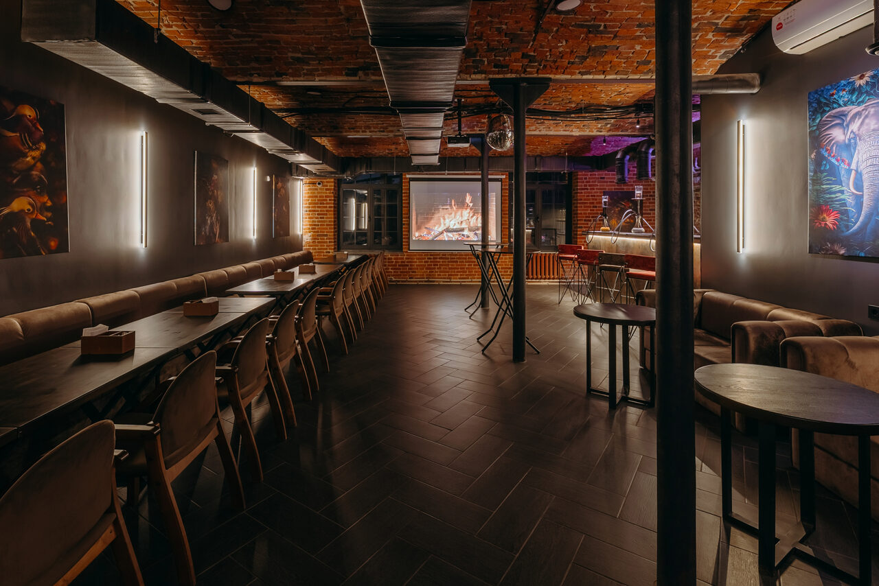 Арендовать Авторские пространство T&S loft (ти энд си лофт) . Тёмное помещение с кирпичными стенами и черным потолком. Длинный стол и стулья слева, диван и столики справа. Проектор с изображением камина на дальней стене. Вертикальные светильники на стенах, картины и кондиционер.