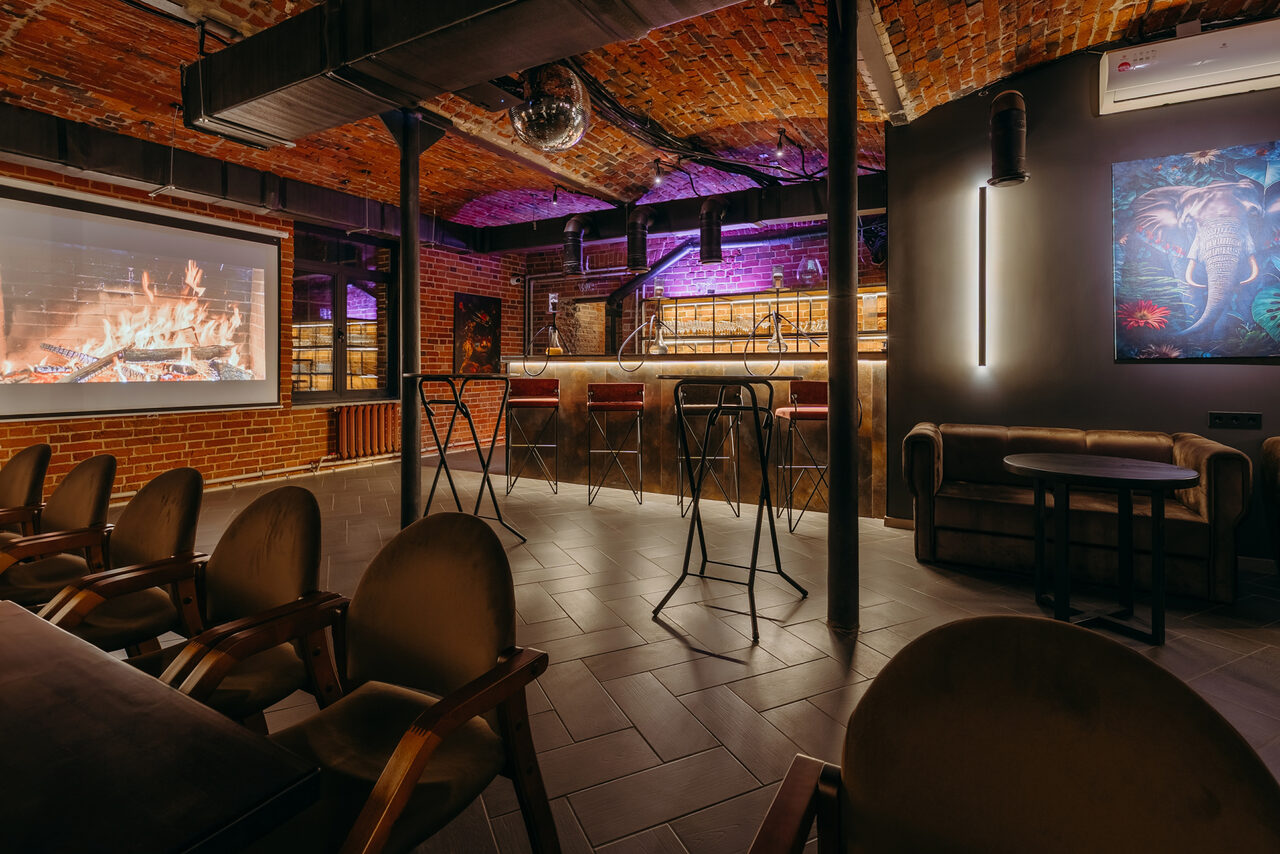 Арендовать Авторские пространство T&S loft (ти энд си лофт) . Лофт-пространство с кирпичными стенами и арками, барная стойка с высокими стульями, проектор с изображением камина, мягкие кресла, неоновая подсветка, настенный кондиционер, декоративная картина, напольное покрытие из серой плитки.