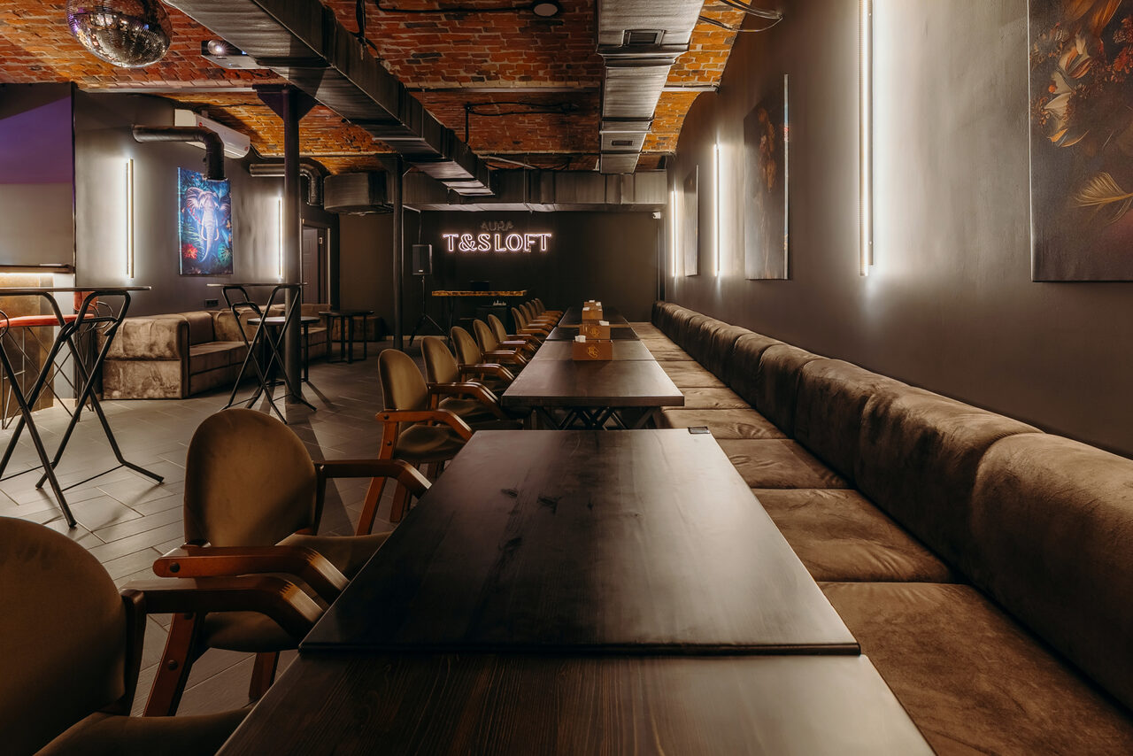 Арендовать Авторские пространство T&S loft (ти энд си лофт) . Лофт-пространство с кирпичным потолком. Темные деревянные столы, стулья с мягкими спинками, длинный диван вдоль стены. Настенные неоновые лампы и картины. Столики для двоих с высокими стульями. Надпись "T&S LOFT" на задней стене.