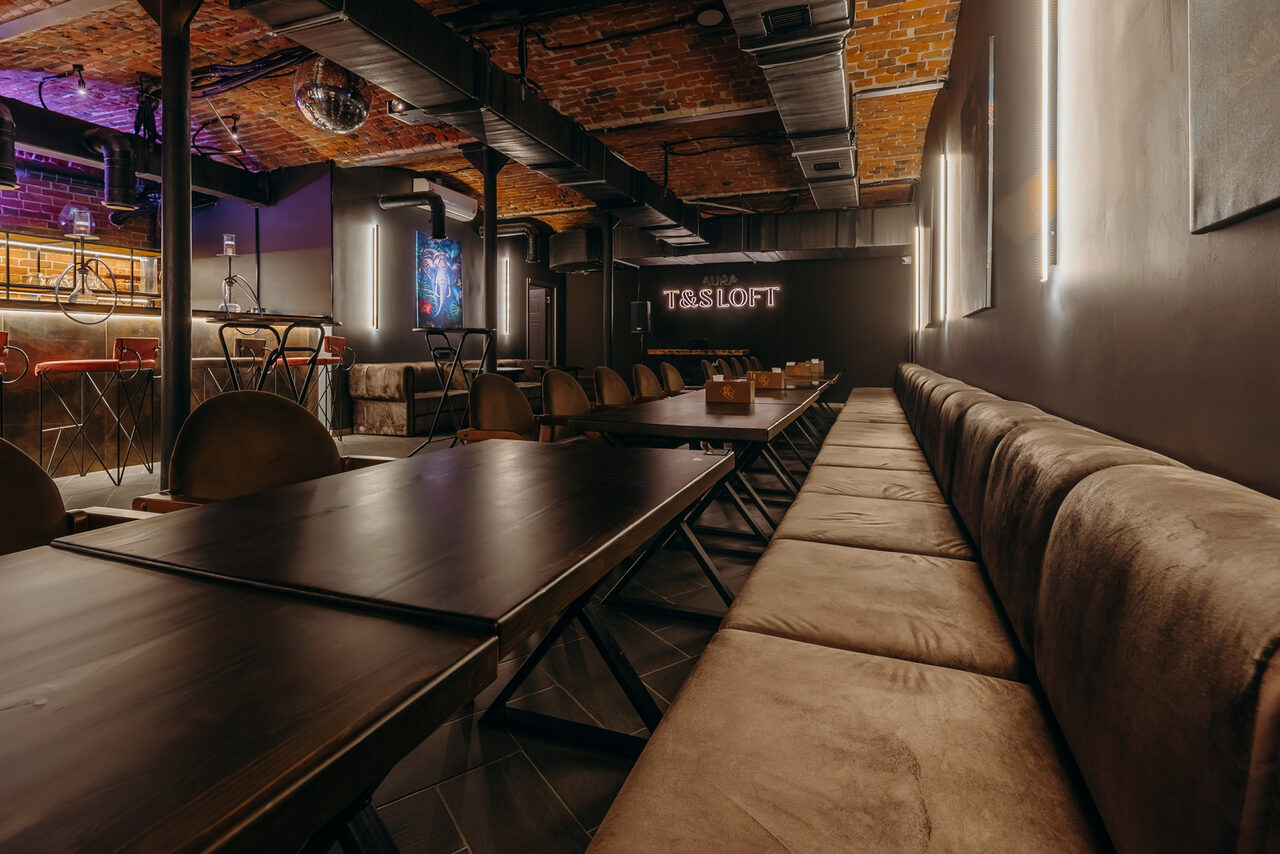 Арендовать Авторские пространство T&S loft (ти энд си лофт) . Лофт-пространство с кирпичной стеной и потолком. Длинный стол с темными столешницами и рядом стульев. Сбоку мягкий диван вдоль стены. Барная стойка с высокими стульями на заднем плане. Неоновая надпись на стене. Подвесное освещение и декоративные элементы.