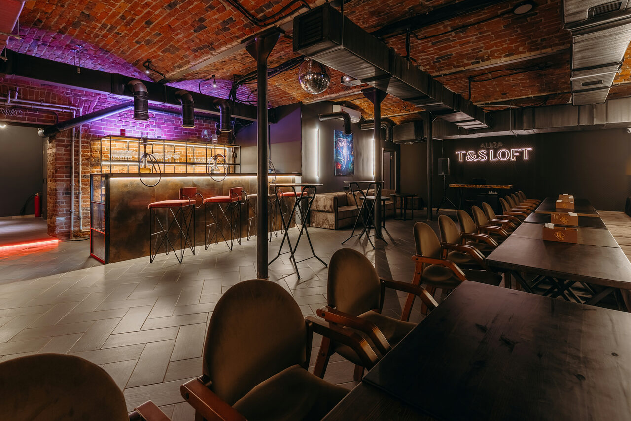 Арендовать Авторские пространство T&S loft (ти энд си лофт) . Лофт-пространство с кирпичными стенами и потолком, деревянной мебелью, длинными столами и стульями. Есть барная стойка с металлическими элементами, высокие барные стулья, неоновая вывеска на стене, мягкое освещение. Потолок с вентиляционными трубами.