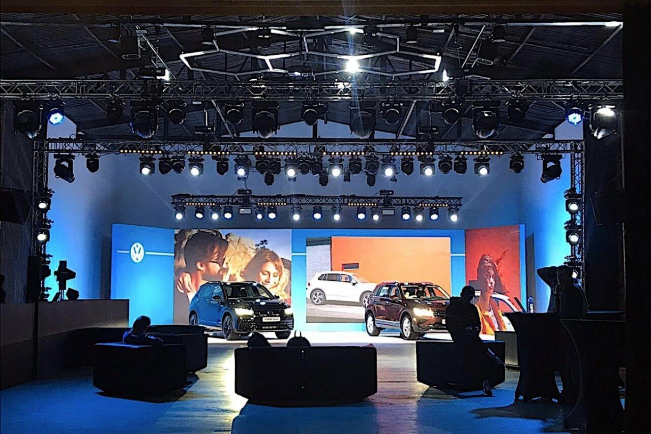 Арендовать Studio Hall(Студио Холл). Сцена с двумя автомобилями, освещённая яркими прожекторами и окружённая синим светом. В помещении видны конструкции с тросами и софитами. На заднем плане крупные экраны с изображениями автомобилей. Впереди мягкие чёрные кресла.