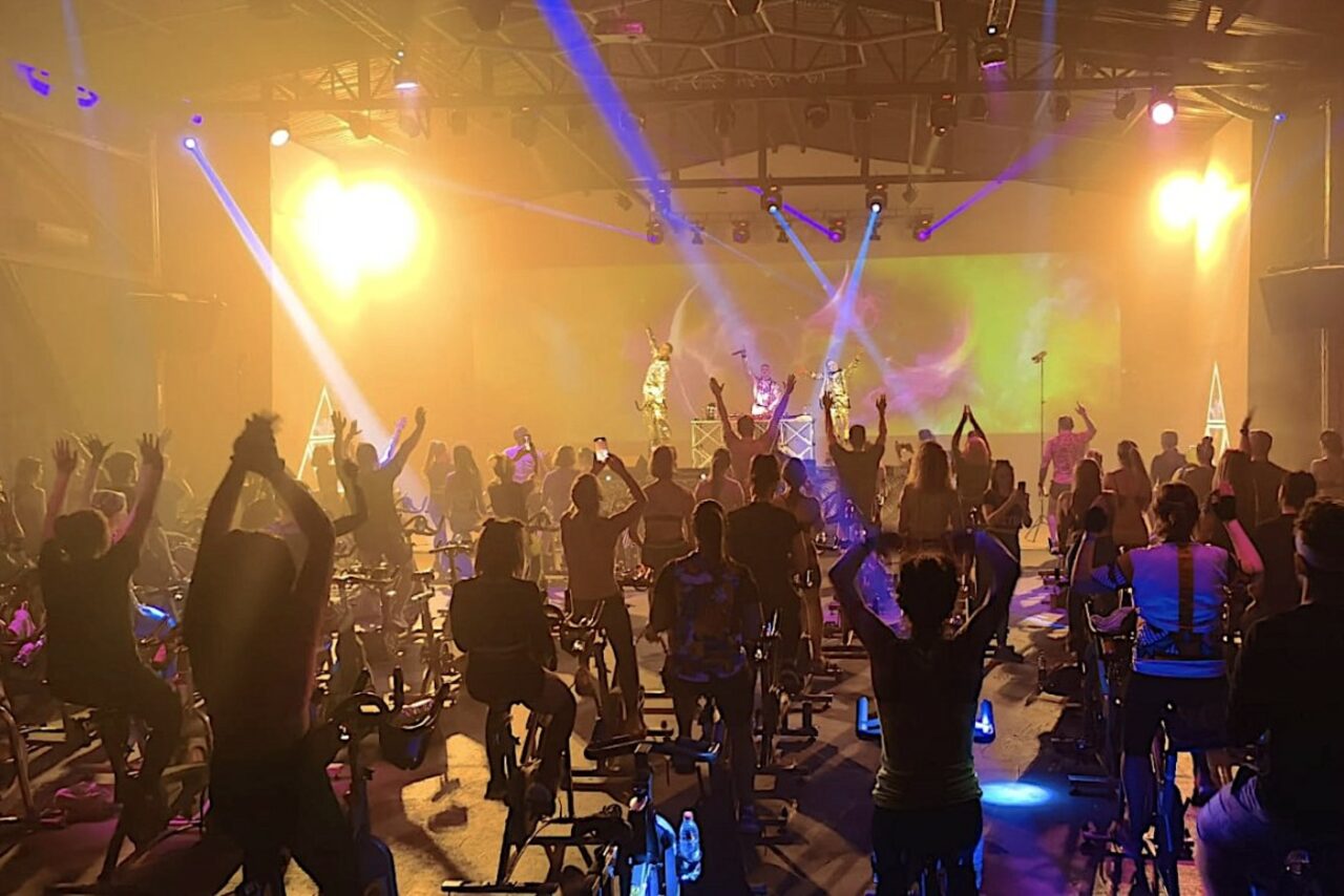 Арендовать Studio Hall(Студио Холл). Атмосферное помещение для групповых тренировок, расставлены велотренажеры, много людей занимается. Яркое освещение, световые эффекты с прожекторами, виден экран. Потолок с видимыми балками, полы тёмного цвета, индустриальный стиль.