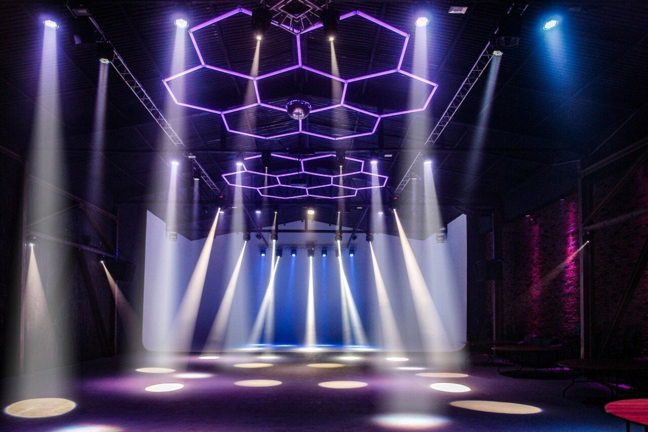 Арендовать Studio Hall(Студио Холл). Просторное помещение с черным потолком и кирпичными стенами. Видна сцена с белыми занавесями. Световое оборудование создает разноцветные световые пятна и линии, включая сиреневые и синие оттенки. Потолок декорирован шестигранниками.