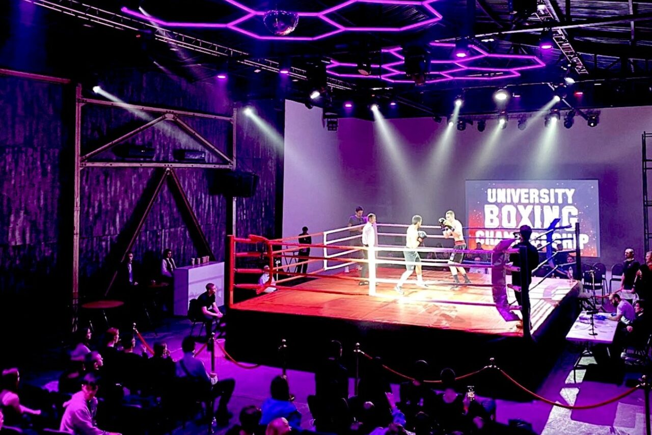 Арендовать Studio Hall(Студио Холл). Изображение показывает спортивное пространство с боксерским рингом в центре. Вокруг ринга собрались зрители. Зал освещен фиолетовыми и белыми прожекторами. На стене экрана видна надпись "University Boxing Championship".
