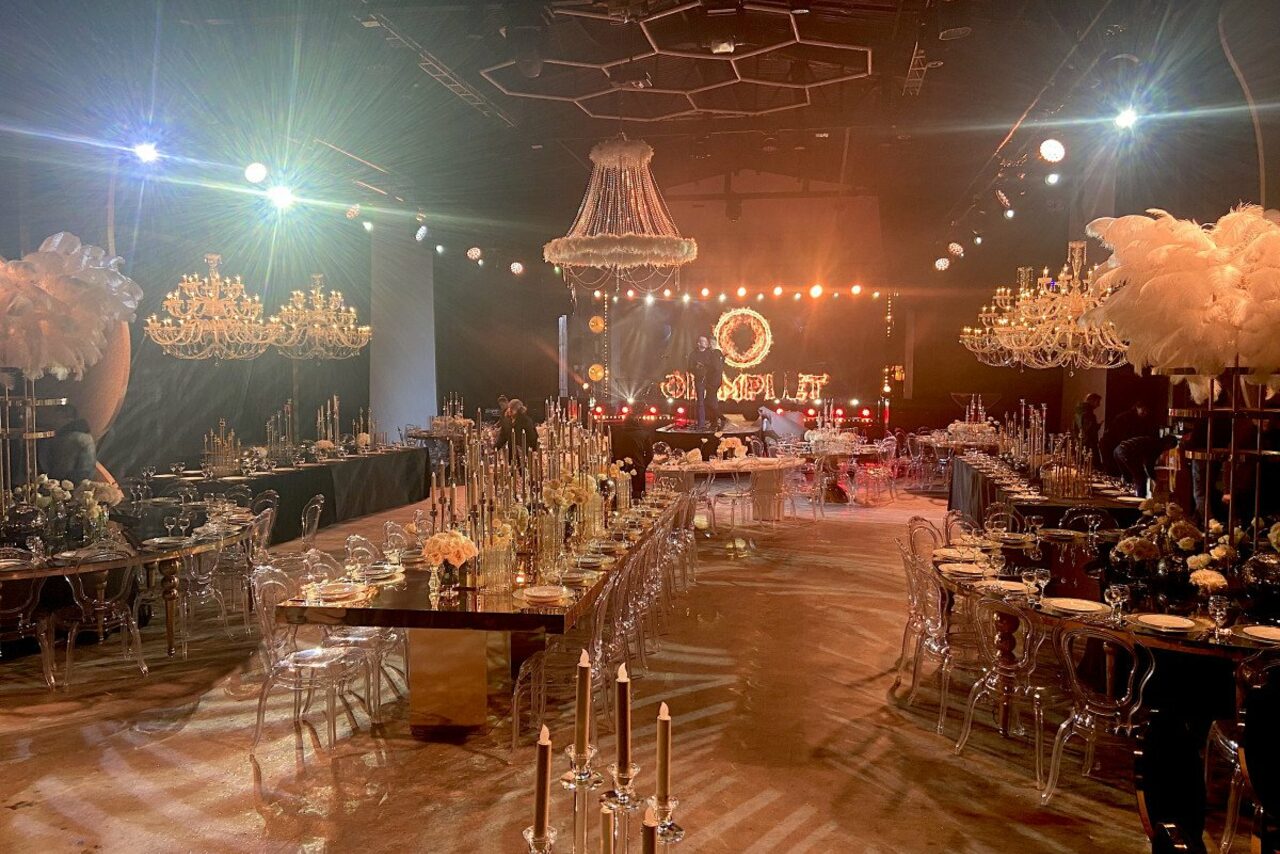 Арендовать Studio Hall(Студио Холл). Большой банкетный зал с длинными столами и прозрачными стульями. На столах украшения и свечи. Потолочные люстры и перьевые декорации. Зал освещён тёплыми лампами, светильники на потолке. Цветовая гамма — золотисто-коричневая.
