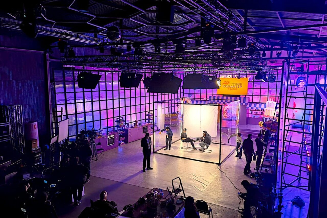 Арендовать Studio Hall(Студио Холл). Просторное помещение, напоминающее студию, с высокими потолками и сетчатой конструкцией. В центре — ярко освещенная зона с кубической конструкцией и людьми. Задний план с крупными стеклянными панелями и цветным освещением. Оборудование и провода видны по периметру.