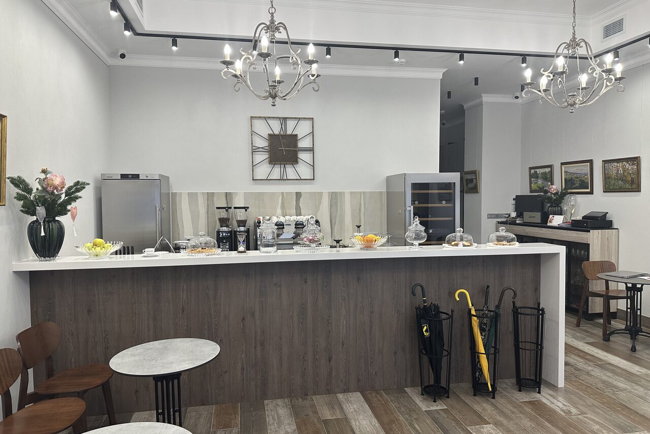 Арендовать Арт Холл. Кафе с барной стойкой из дерева, белой столешницей. На стойке кофемашина, вазы с фруктами и десертами. Сзади холодильник и шкаф. Светильники на потолке, картины на стенах. Деревянные пол и стулья, столы с мраморными столешницами. Зонты за стойкой.