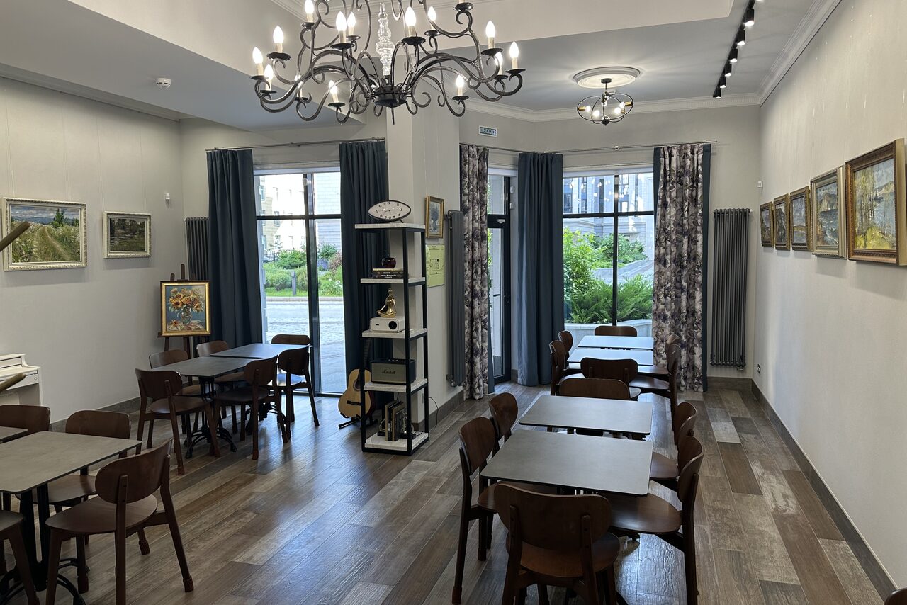 Арендовать Арт Холл. Небольшое помещение с кафейными столами и стульями, стены украшены картинами. На полу деревянный ламинат, освещение от современных и классических люстр. Большие окна с плотными шторами. Справа книжная полка с декорациями.