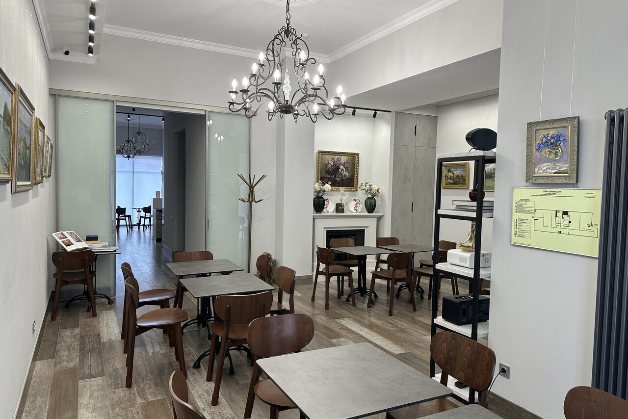 Арендовать Арт Холл. Комната с деревянными столами и стульями, белые стены с картинами, подвесная люстра. Стеклянная дверь ведет в соседнее помещение. Пол из светло-коричневого дерева. В углу стоит вешалка, у стены камин. На стене план помещения.