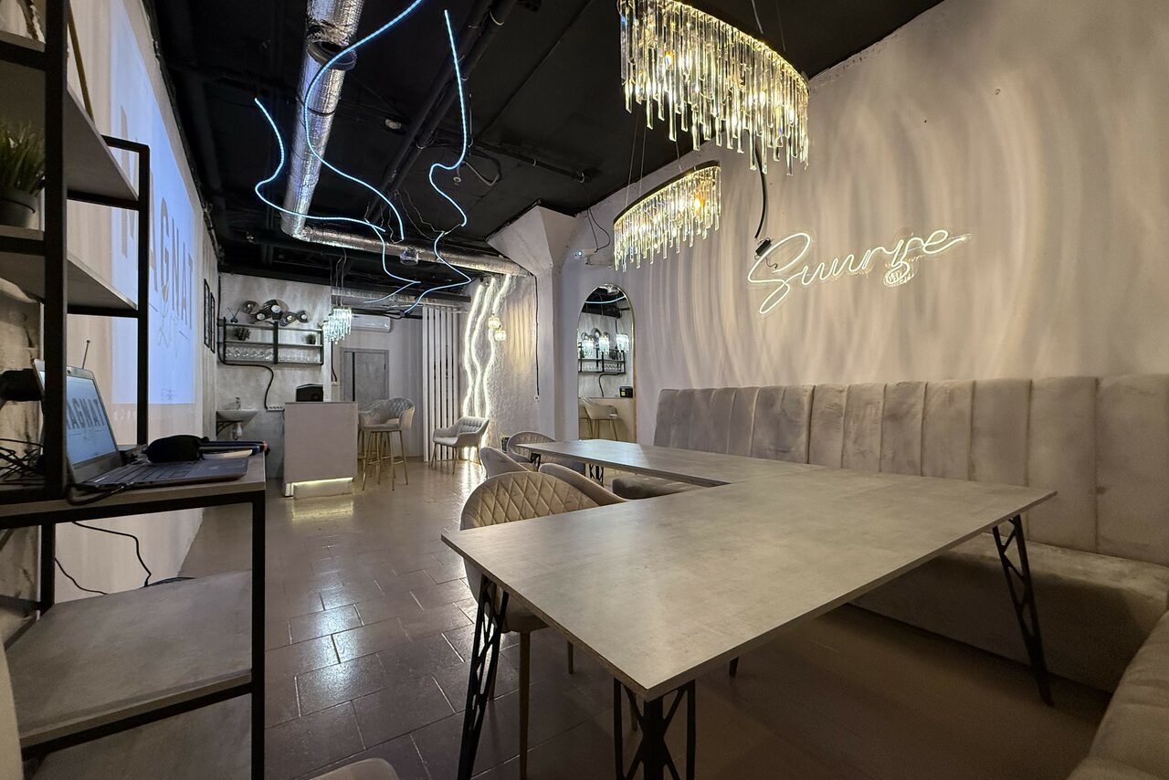 Арендовать Лофт SUNRISE(Восход Солнца). Просторное помещение на 15 гостей. Пространство с длинным столом и стульями, мягкими стеновыми сидениями. На стене неоновая вывеска. Потолок с видимыми коммуникациями, декоративное освещение. Пол из плитки. Современный интерьер.