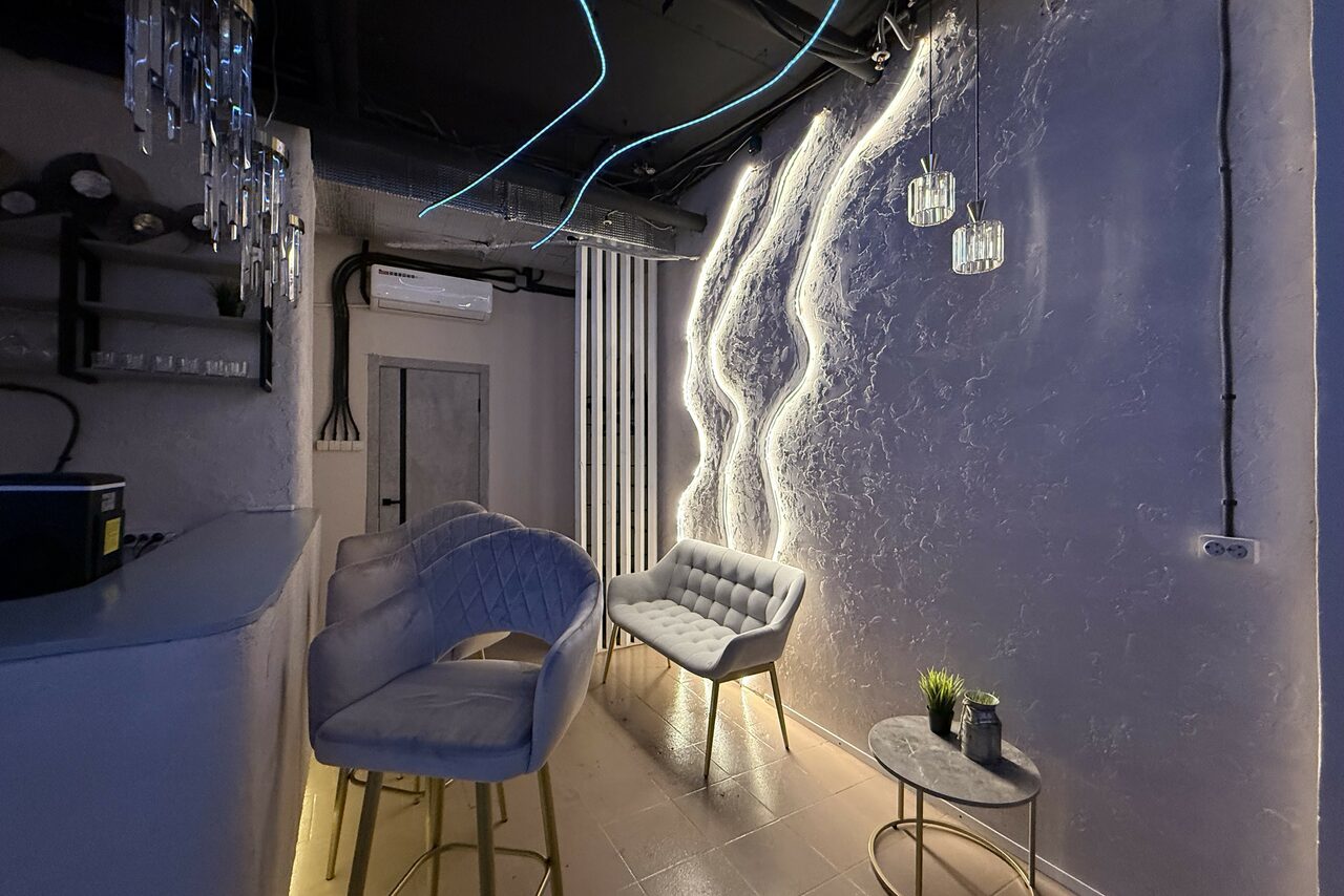 Арендовать Лофт SUNRISE(Восход Солнца). Просторное помещение на 15 гостей. Современное помещение с барной стойкой и мягкими стульями. Стены с акцентным освещением и фактурной отделкой, подвесные стеклянные светильники, обивка мебели в серых тонах, стеклянный столик с декором, бежевый пол, кондиционер и видимые трубы на потолке.