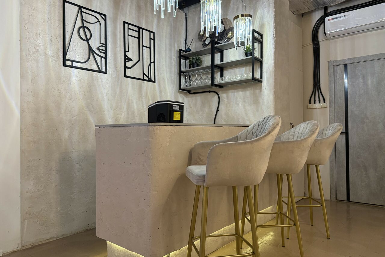 Арендовать Лофт SUNRISE(Восход Солнца). Просторное помещение на 15 гостей. Барная стойка с тремя стульями на золотистых ножках. Стена из светлого материала с геометрическими рисунками. Полки с декором и бокалами. Подвесное освещение с лампами. Настенный кондиционер в верхнем углу. Светлый пол.
