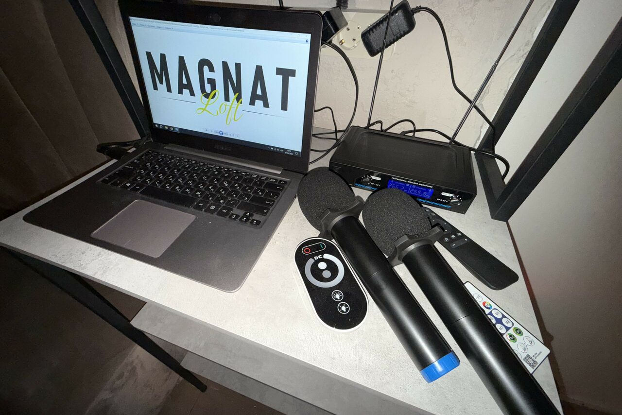 Арендовать Лофт SUNRISE(Восход Солнца). Просторное помещение на 15 гостей. Ноутбук на светлом столе, открыт экран с текстом "MAGNAT Loft". Рядом два микрофона, пульт управления, дистанционный пульт и кнопка. На стене черные провода и устройство с синим дисплеем. Стена светлая.