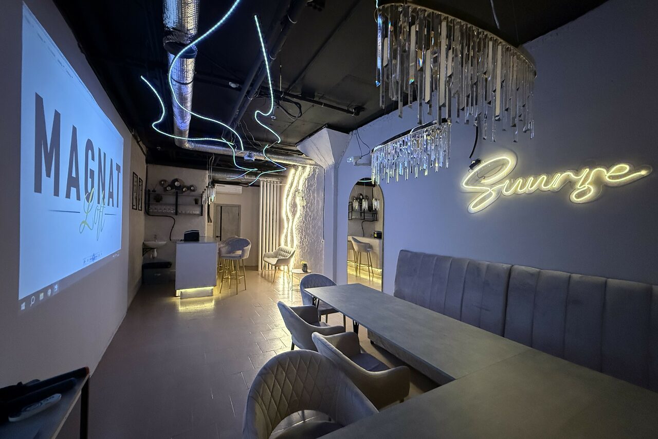 Арендовать Лофт SUNRISE(Восход Солнца). Просторное помещение на 15 гостей. Изображение современного интерьера с баром и зоной отдыха. На стене неоновая надпись, мягкие диваны и кресла вокруг длинных столов. Наверху свето-диодные ленты и стеклянные подвески. На экране проекция. Тёмный потолок с видимыми трубами.