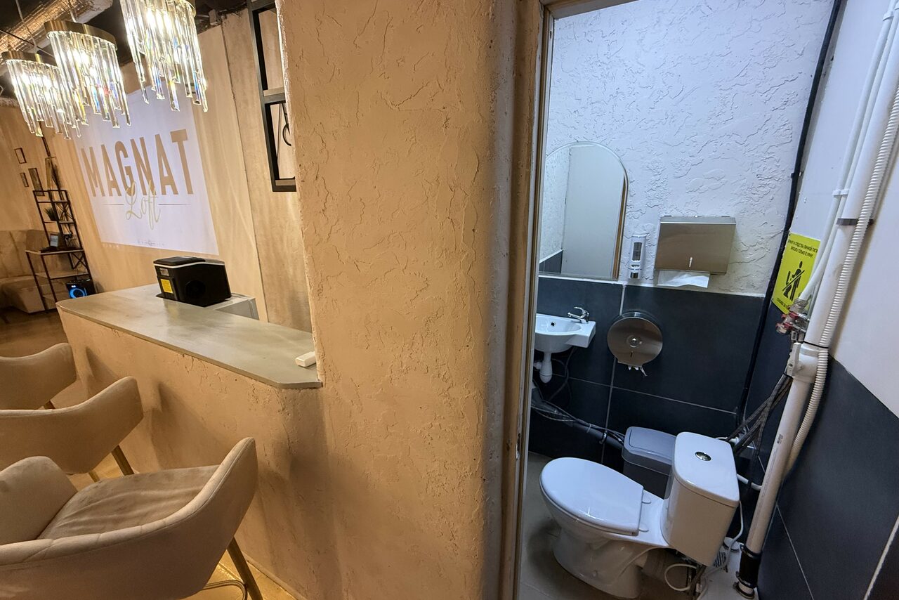 Арендовать Лофт SUNRISE(Восход Солнца). Просторное помещение на 15 гостей. Барная стойка примыкает к стене, рядом зона с туалетом. На баре несколько стульев, светлое освещение. Туалет оборудован унитазом, раковиной, зеркалом. Стены с текстурной отделкой, контраст серого и бежевого цветов. Надпись на стене: MAGNAT.