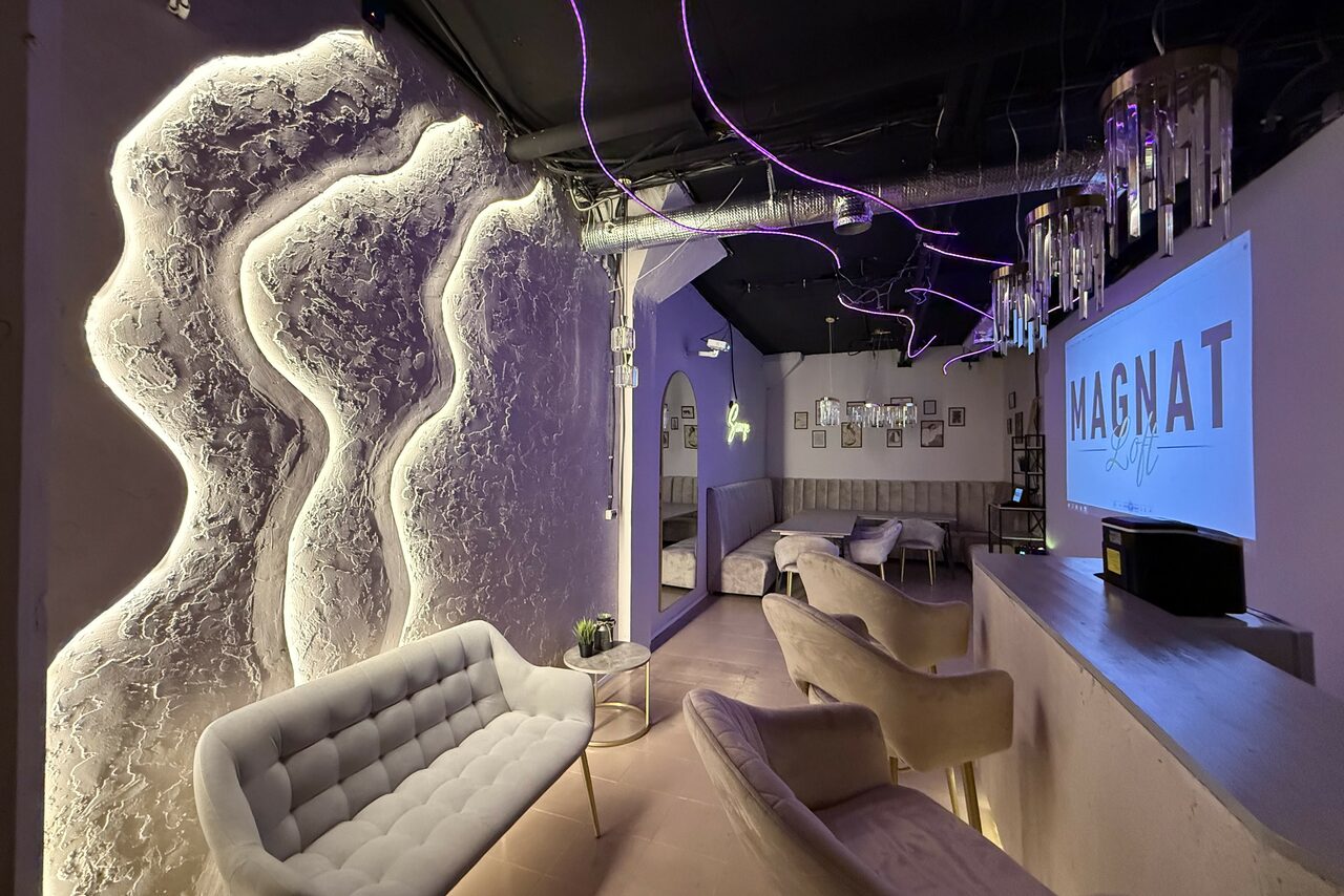 Арендовать Лофт SUNRISE(Восход Солнца). Просторное помещение на 15 гостей. Современное помещение с приглушенным освещением, мягкими креслами и диванами, декоративной подсветкой на стенах. На стене знак "MAGNAT". Видно барную стойку, подвесные светильники, трубы на потолке, картины на стене.
